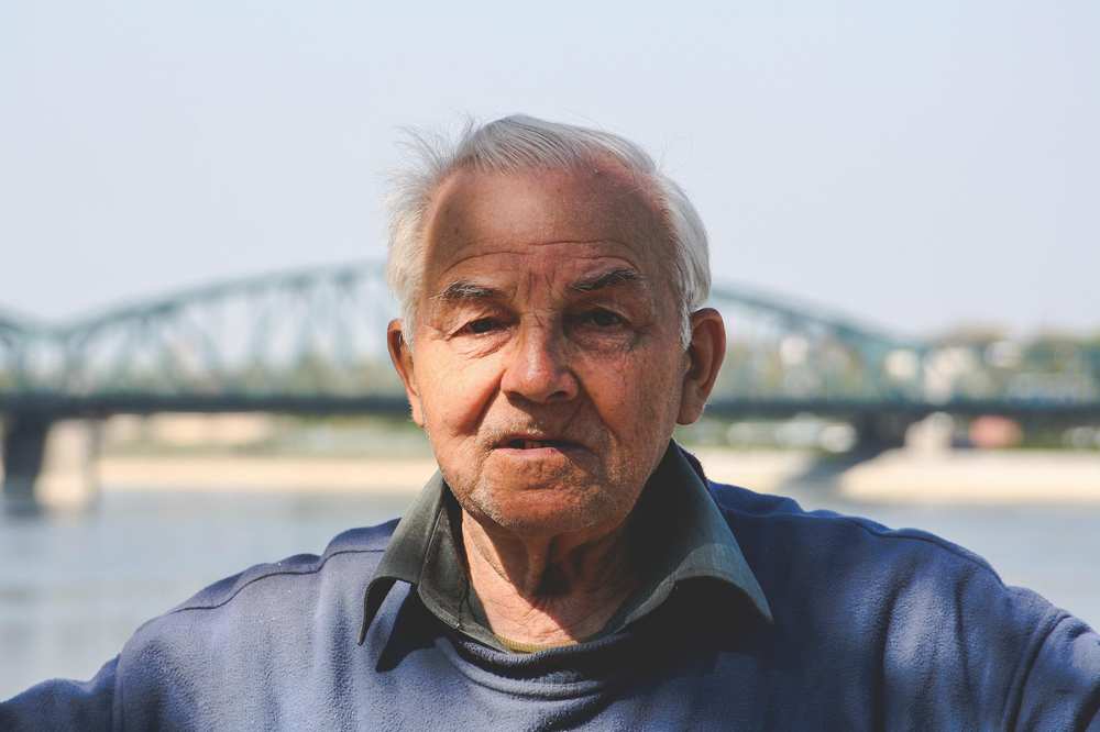 Ein älterer Mann. Im Hintergrund ist eine Brücke über einen Fluss zu sehen. 