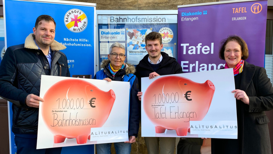 Im Hintergrund hängen Plakate mit den Logos von Tafel Erlangen und der Bahnhofsmission. Im Vorderund stehen zwei Männer und zwei Frauen mit symbolischen Schecks.