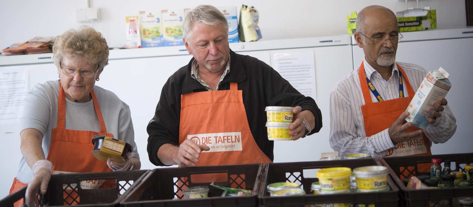 Drei Mitarbeitende mit organgenen Tafelschürzen sortieren Lebensmittel in Plastikboxen.