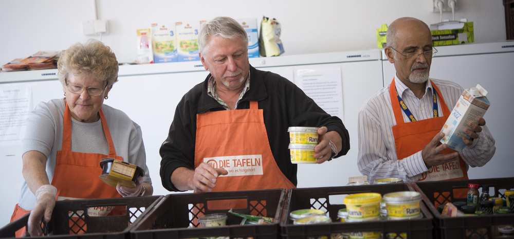 Drei Mitarbeitende mit organgenen Tafelschürzen sortieren Lebensmittel in Plastikboxen.