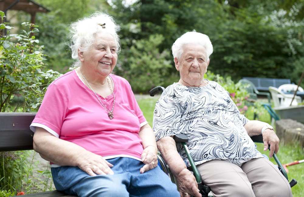 Zwei ältee Frauen sitzen im Garten. Im Hintergrund stehen Blumenkästen und eine Bank. Der Rasen und die Bäume werden von der Sonne beschienen.